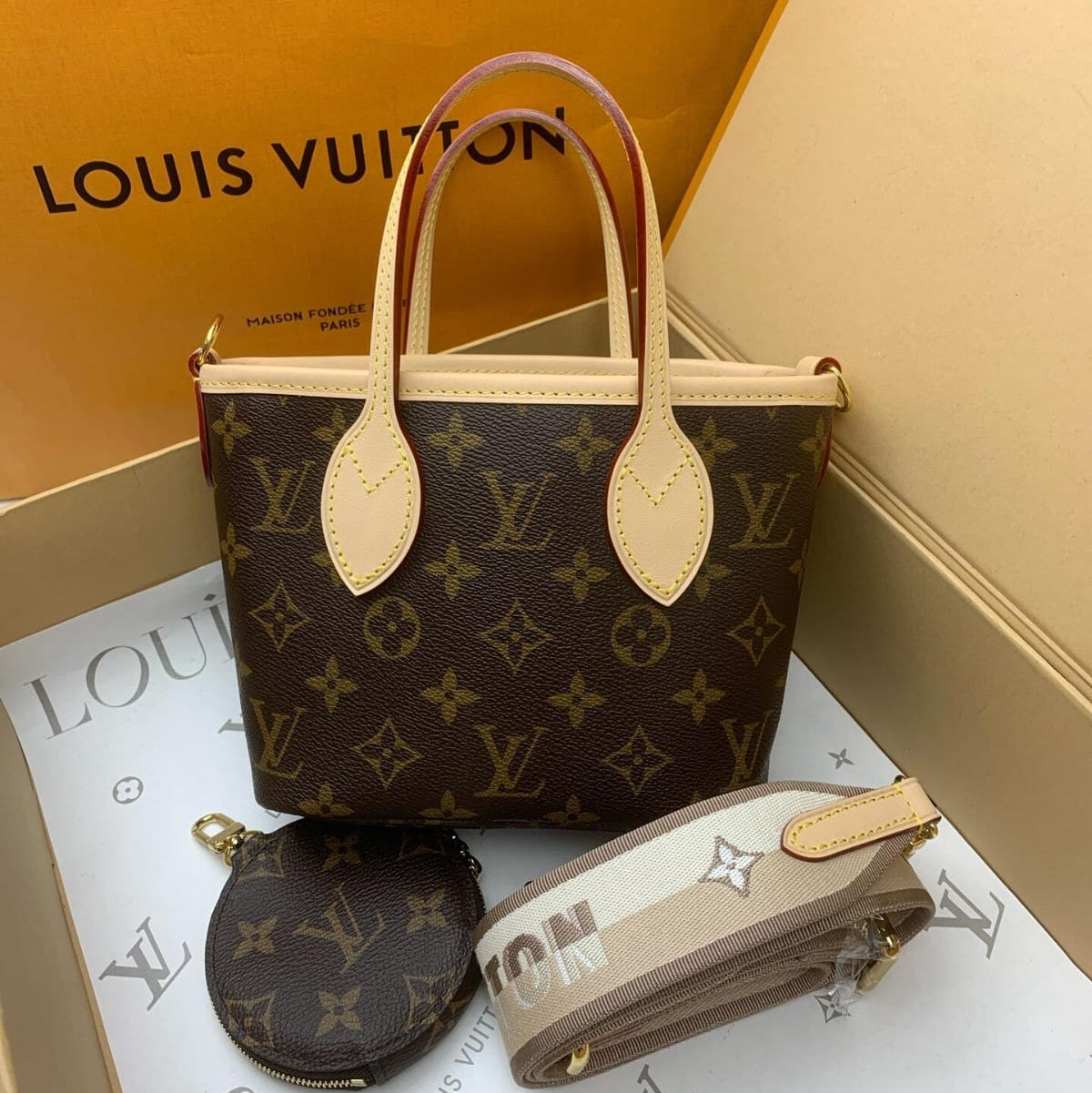 Louis Vuitton Neverfull tote bag in brown monogram canvas with beige leather handles, shown with a matching coin pouch and a striped shoulder strap inside an open Louis Vuitton box.