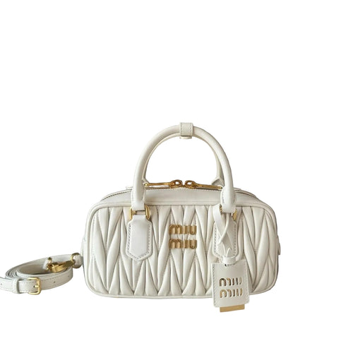 A white Miu Miu quilted leather handbag with gold hardware, featuring dual top handles, a detachable shoulder strap, and a hanging Miu Miu tag, displayed on a light surface against a neutral taupe background.