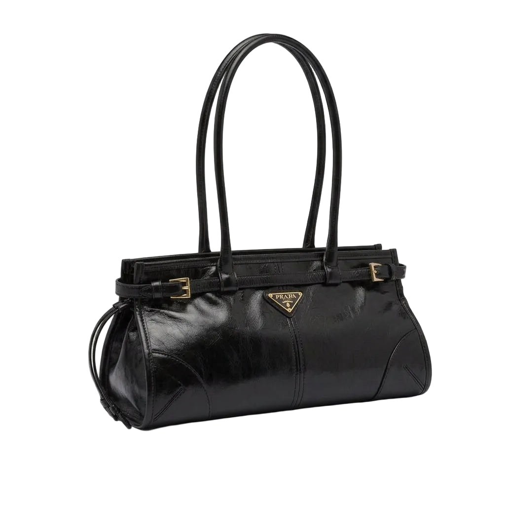 A black Prada leather handbag displayed on a dark table with its white dust bag, shopping bag, and strap laid out in front. The Prada logo is visible on the bag and shopping bag.