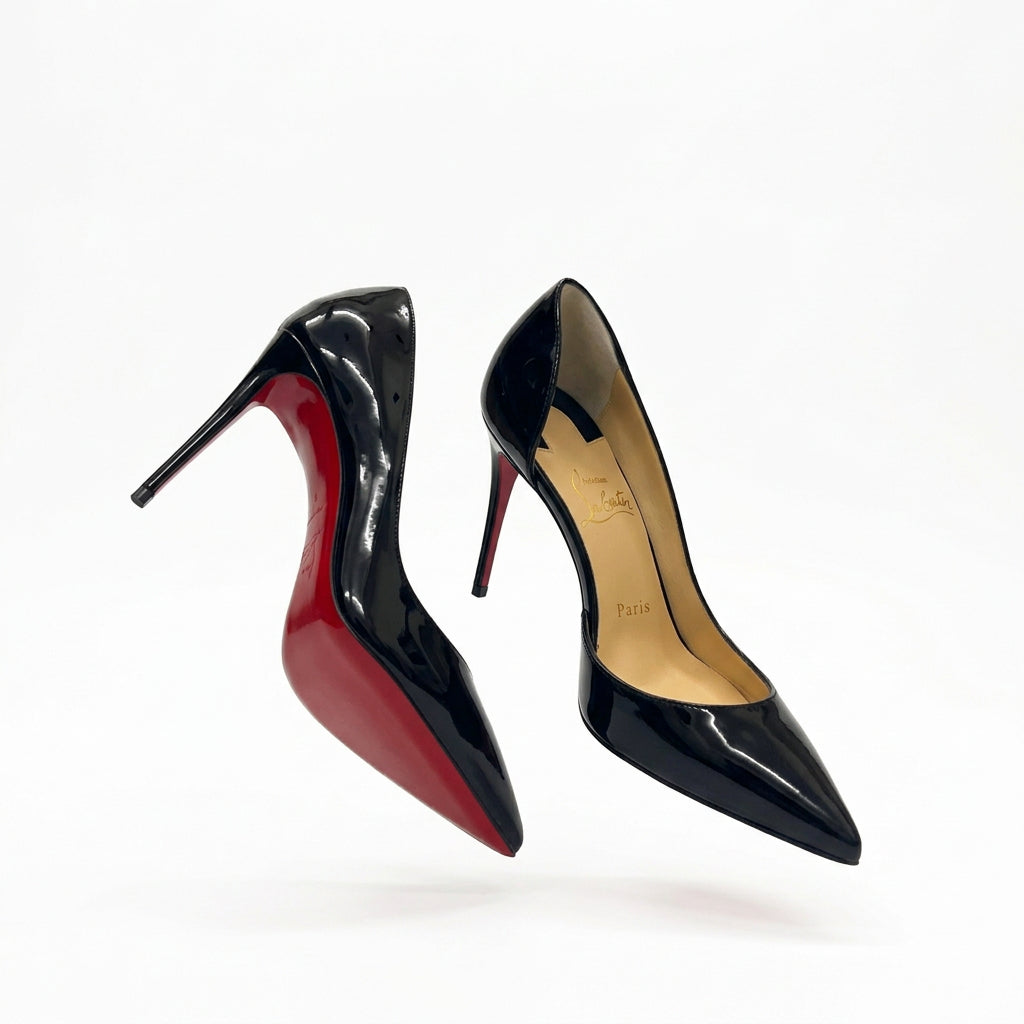 A pair of Christian Louboutin black patent leather pointed-toe pumps with their signature red soles, presented in their white branded dust bag and tissue paper inside the shoebox, with the cardboard box visible in the background on a wooden floor.
