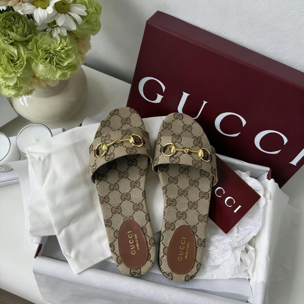 Gucci GG Supreme slide sandals in a white box with white tissue paper, a burgundy Gucci shoebox, and a white Gucci dust bag, styled next to a vase of flowers.