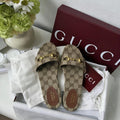Gucci GG Supreme slide sandals in a white box with white tissue paper, a burgundy Gucci shoebox, and a white Gucci dust bag, styled next to a vase of flowers.