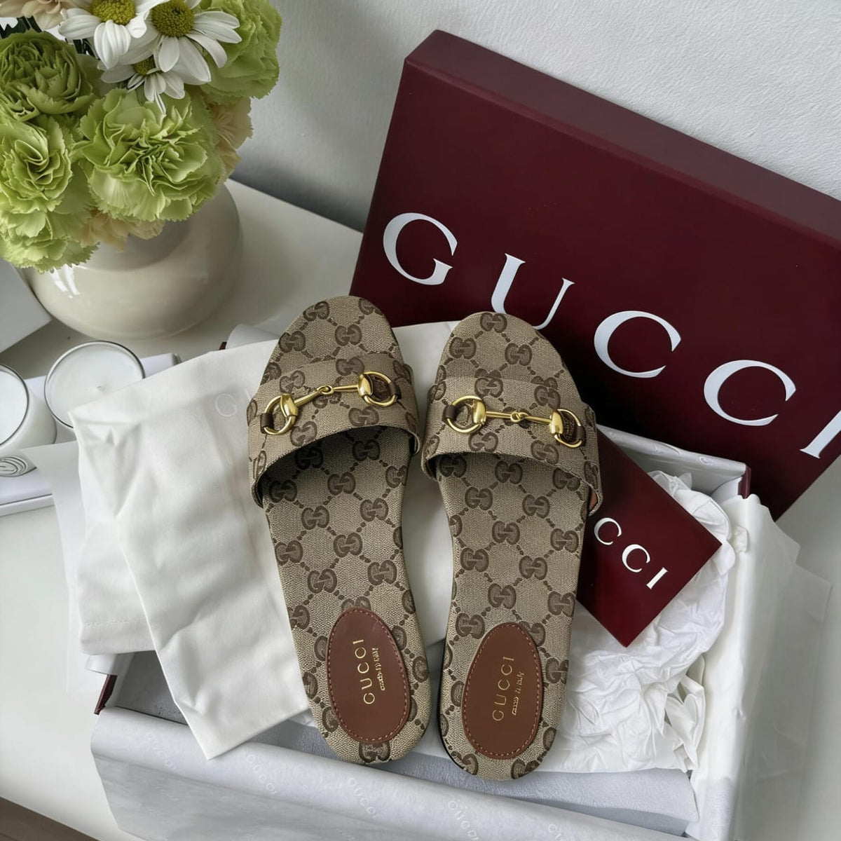 Gucci GG Supreme slide sandals in a white box with white tissue paper, a burgundy Gucci shoebox, and a white Gucci dust bag, styled next to a vase of flowers.