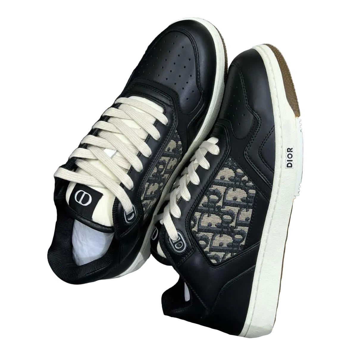 A pair of Dior low-top sneakers in black with beige and white patterned side panels, cream laces, and a white midsole, displayed in a shoebox with a navy blue Dior dust bag.