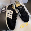 A pair of Dior low-top sneakers in black suede and canvas with white side stripes and 'Dior' branding, displayed on a white Dior shoebox with a Dior perfume bottle in the background.