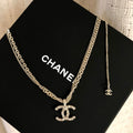 A Chanel necklace with a silver-tone chain and interlocking CC logo pendant encrusted with clear stones, displayed on a black Chanel box against a textured cream fabric background.
