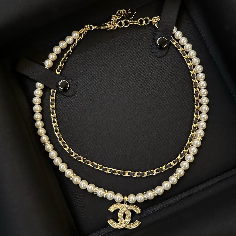 A double-strand Chanel necklace featuring white faux pearls and gold-tone chains, with a gold-tone interlocking CC logo pendant, displayed inside a black presentation box.
